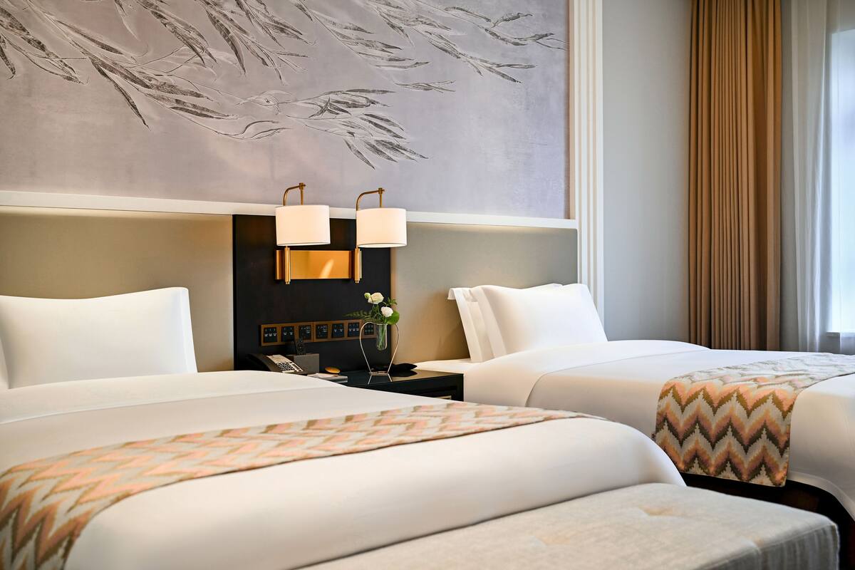 A hotel room with two beds, a large wall mural, a nightstand with lamps, and a view of the outdoors.