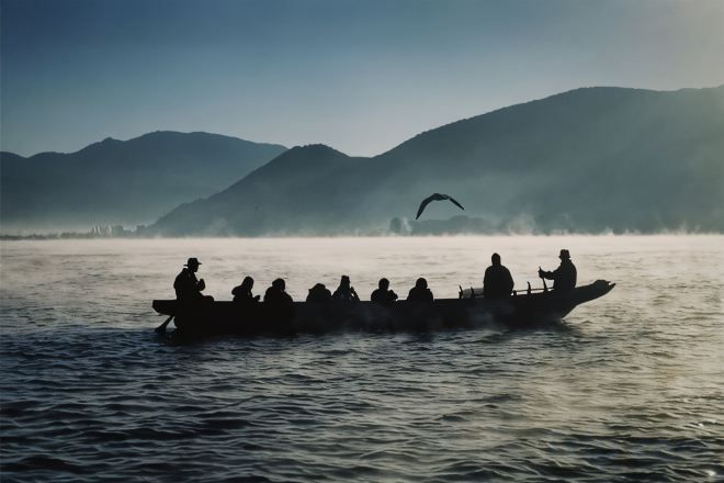 A boat with several people on a calm body of water, surrounded by misty mountains.