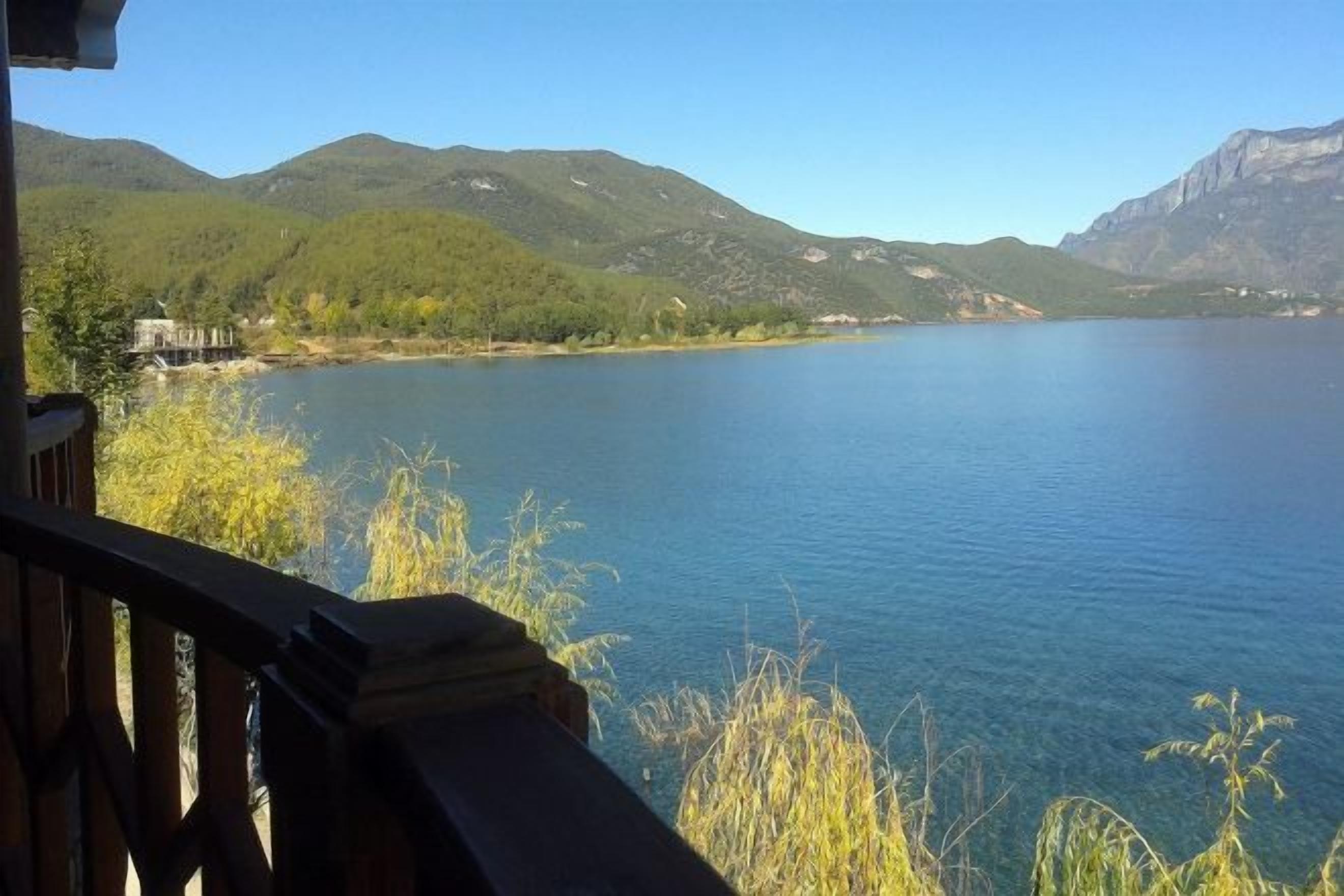 Un lago con montañas al fondo, una terraza de madera y algo de vegetación en primer plano.