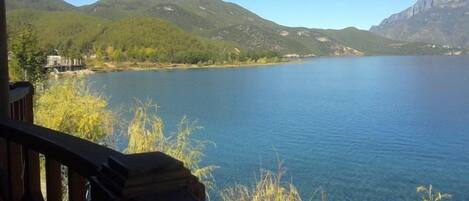 Un lago con montañas al fondo, una terraza de madera y algo de vegetación en primer plano.