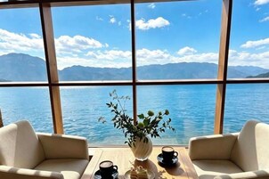 Una habitación con un gran ventanal que da a un cuerpo de agua y montañas, un florero con flores, dos sillones y una mesa con tazas y un plato.