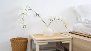 Une petite table en bois sur laquelle est posé un vase blanc contenant des fleurs blanches, un panier en osier et un lit avec une literie blanche.