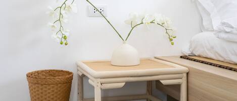 Un tavolino in legno con un vaso bianco pieno di fiori bianchi, un cesto in vimini e un letto con biancheria da letto bianca.