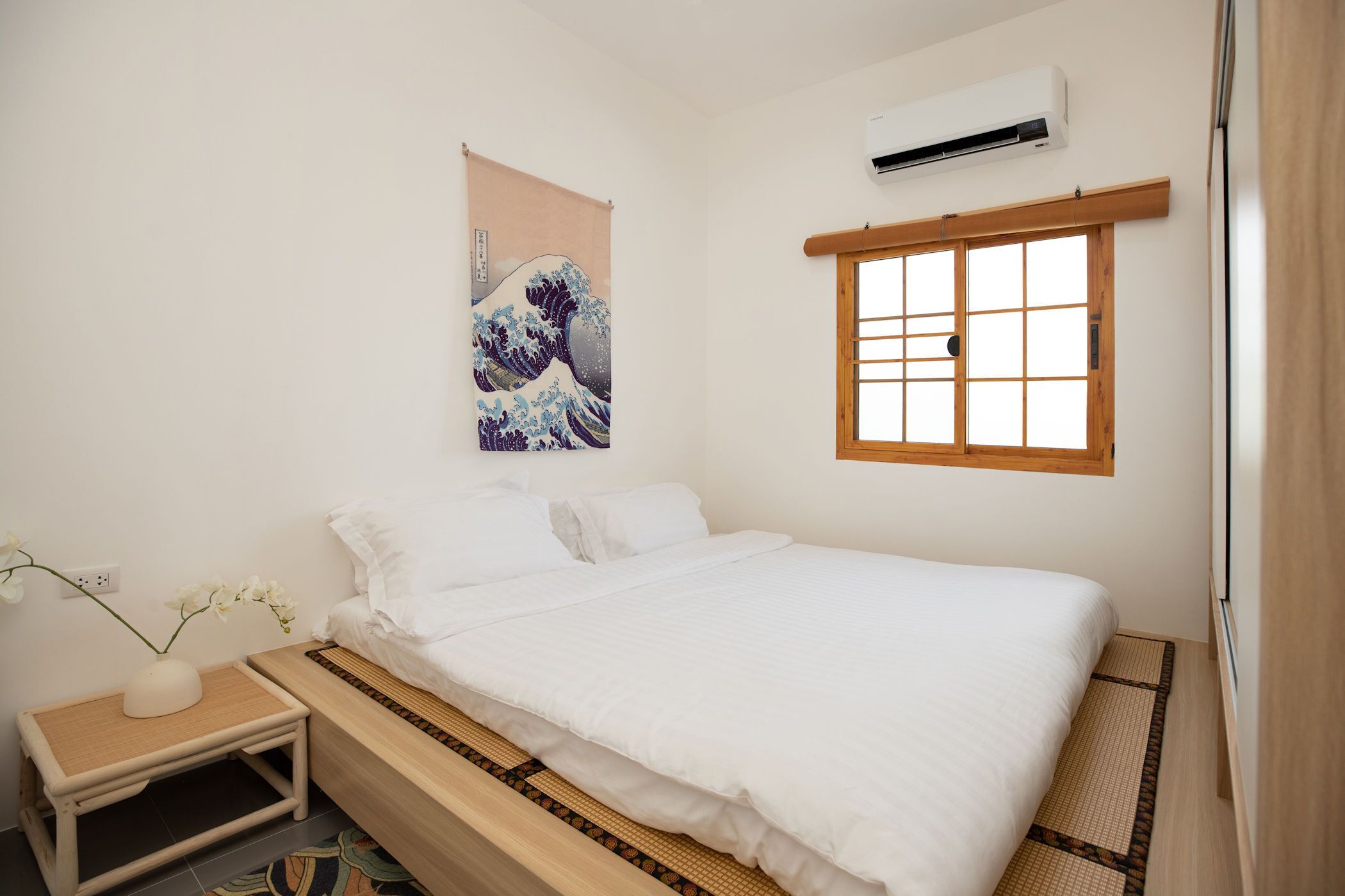 A minimalist bedroom with a bed, a small bedside table, a wall-mounted air conditioner, and a window with wooden frames.