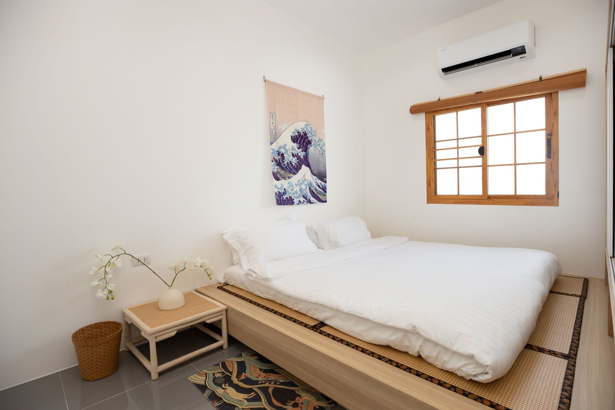 A minimalist Japanese-style bedroom with a bed, a small table with a vase, a woven basket, and a traditional Japanese rug.