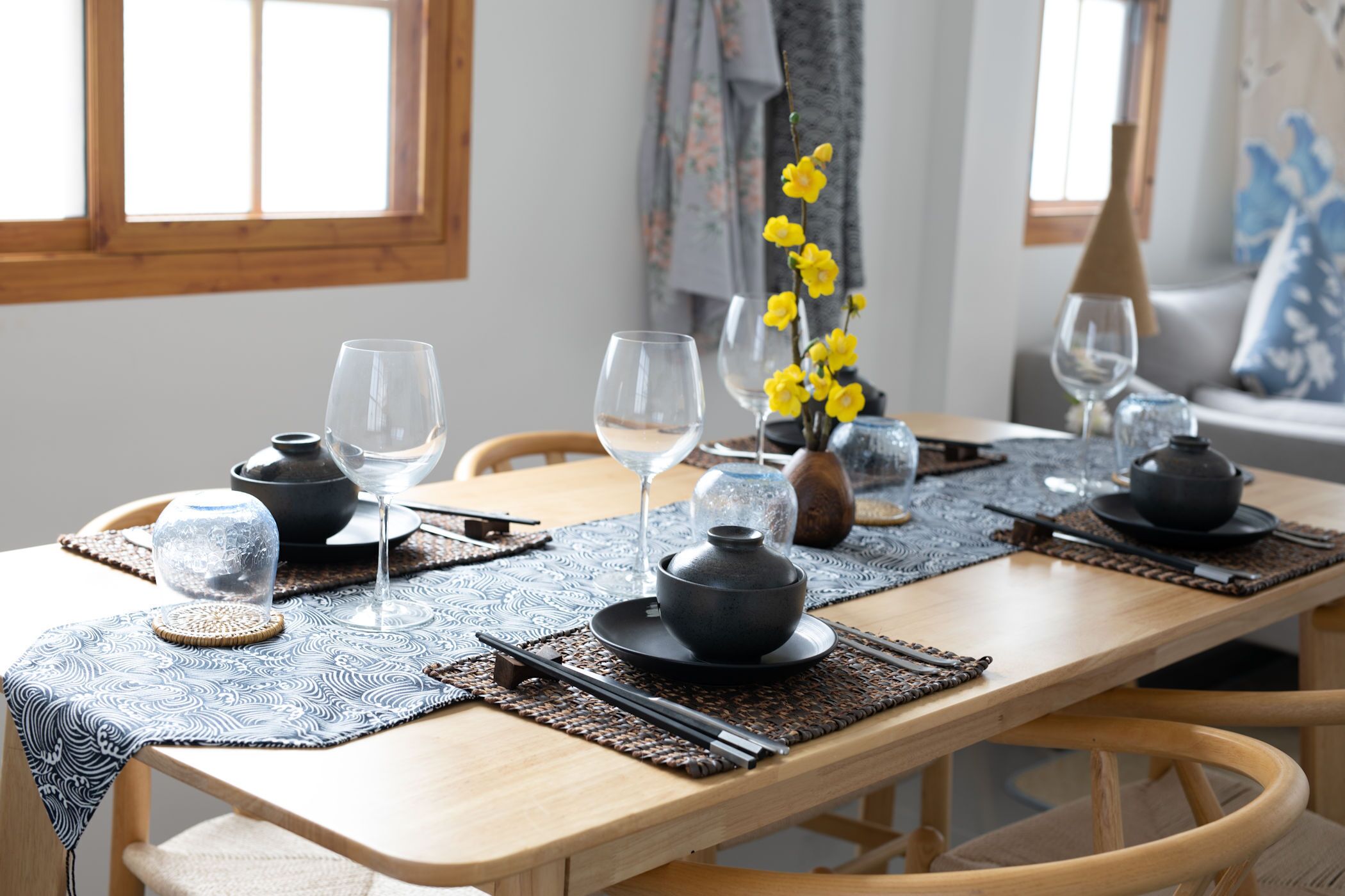 A dining table set with glassware, bowls, and chopsticks, with a woven placemat and a vase of yellow flowers.