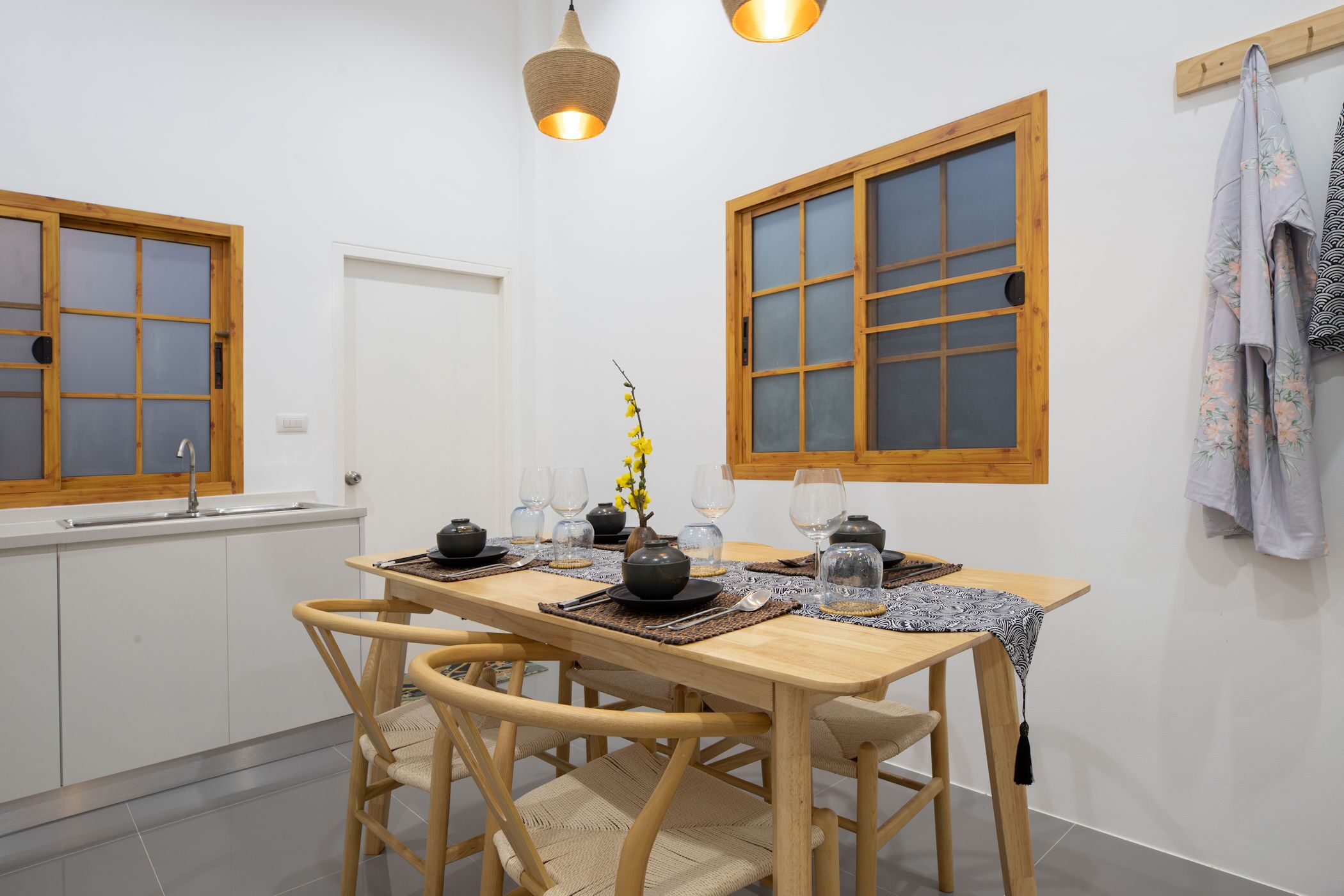 A dining area with a wooden table set for four, two wooden chairs, a vase with yellow flowers, and a window with a wooden frame.