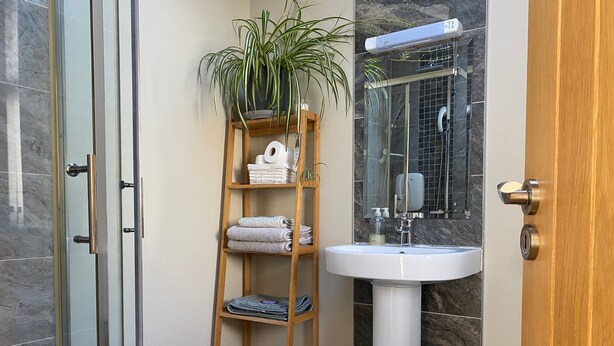 Un baño con mampara de vidrio, un estante de madera con toallas y artículos de aseo, un lavabo blanco y una pared con azulejos de mármol.