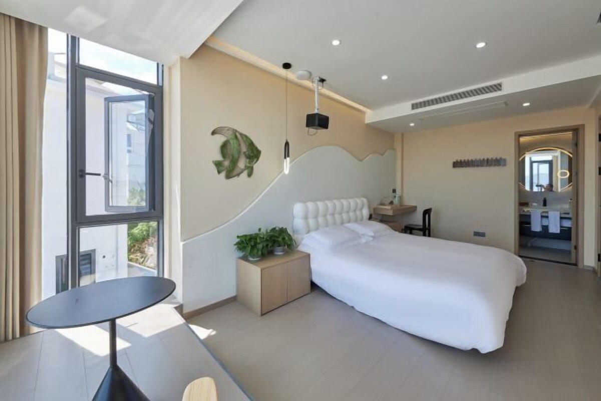 A modern hotel room with a large bed, a small bedside table with a plant, a round table, and a view through a large window.