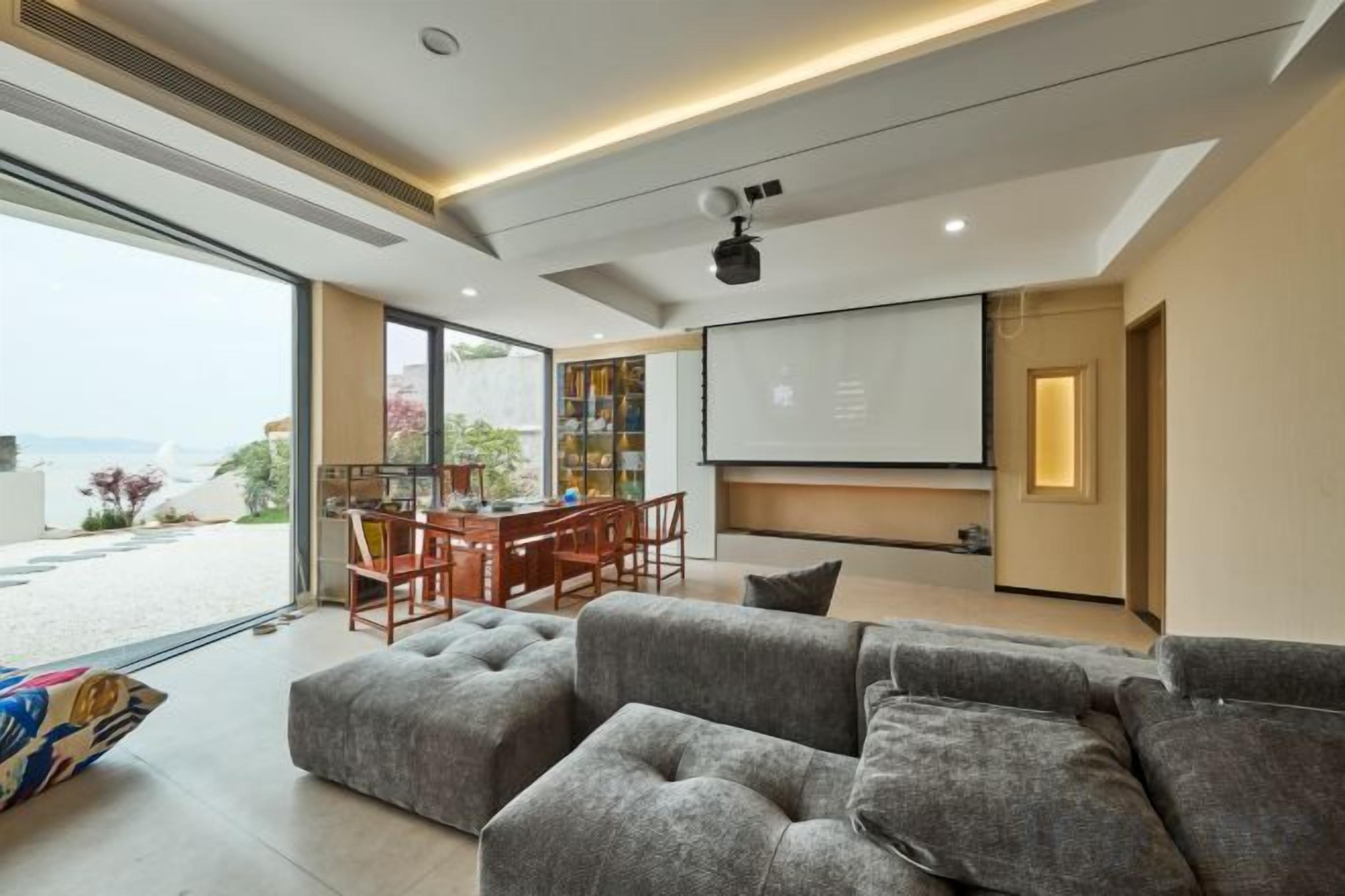 A modern living room with a large gray sectional sofa, a dining area with a wooden table and chairs, and a large flat-screen TV mounted on the wall.