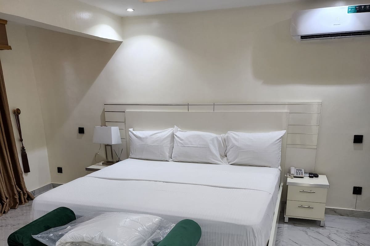 Quarto moderno com uma cama grande, uma poltrona estofada verde, uma mesinha de cabeceira e um ar condicionado.