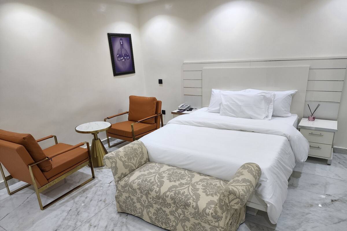 Quarto de hotel com cama, duas poltronas, uma mesinha e um banquinho.