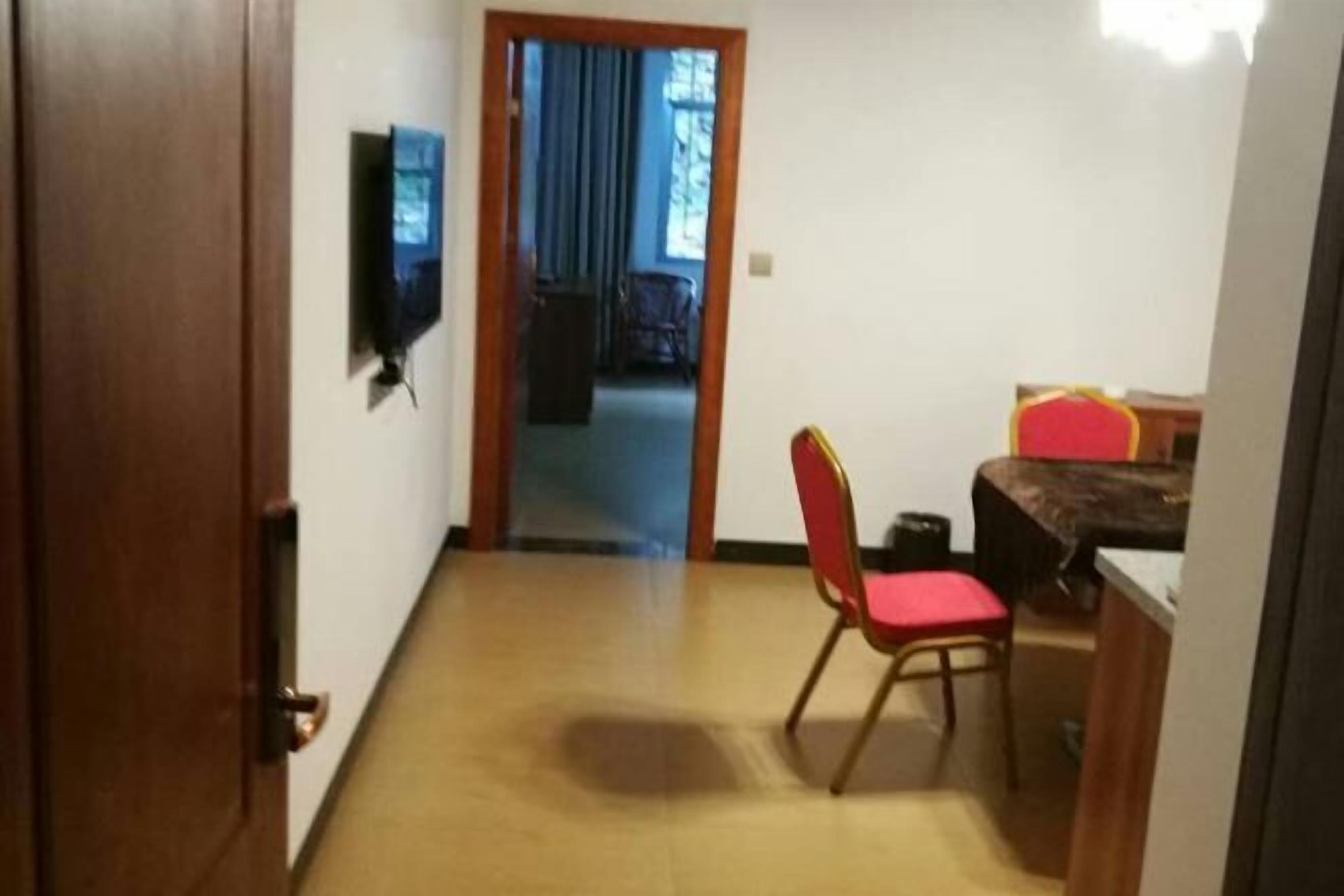 A room with a wooden door, a television mounted on the wall, a dining table with a red chair, and a view of a balcony with blue curtains.