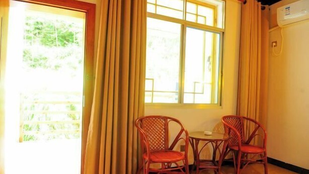 Una habitación con piso de madera, dos sillas de mimbre, una mesita, un ventanal con cortinas y un equipo de aire acondicionado en la pared.