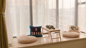 Une pièce chaleureuse avec un coin fenêtre agrémenté de coussins, une petite table avec un service à thé et une vue sur le paysage urbain à travers de fines rideaux.