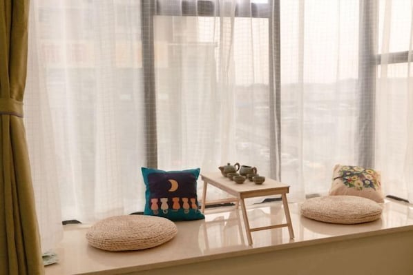 Una habitación acogedora con un asiento junto a la ventana, cojines, una mesita con juegos de té y vistas a la ciudad a través de cortinas translúcidas.