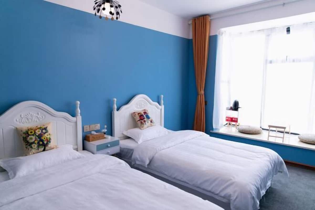 Una habitación con dos camas, una ventana con cortinas, una mesita con lámpara y una lámpara decorativa en el techo.