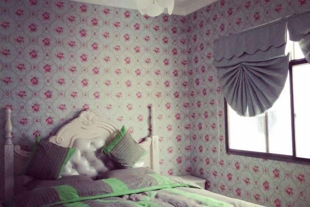 Ein Schlafzimmer mit floraler Tapete, einem Bett mit grün-violetter Bettwäsche und einem Fenster mit grauem Vorhang.