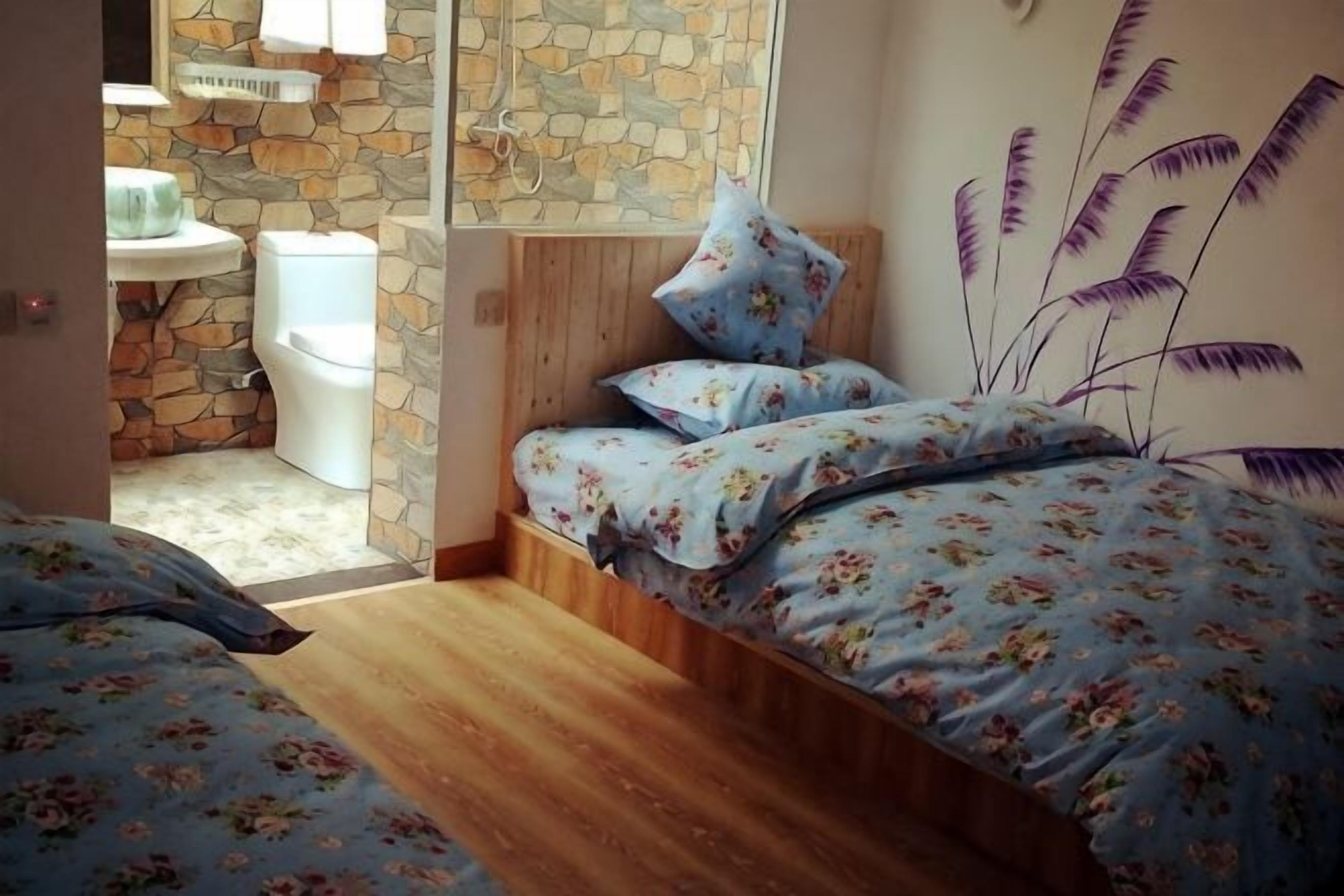 A bedroom with a wooden floor, a bed with floral bedding, a stone wall, and a bathroom with a white toilet.