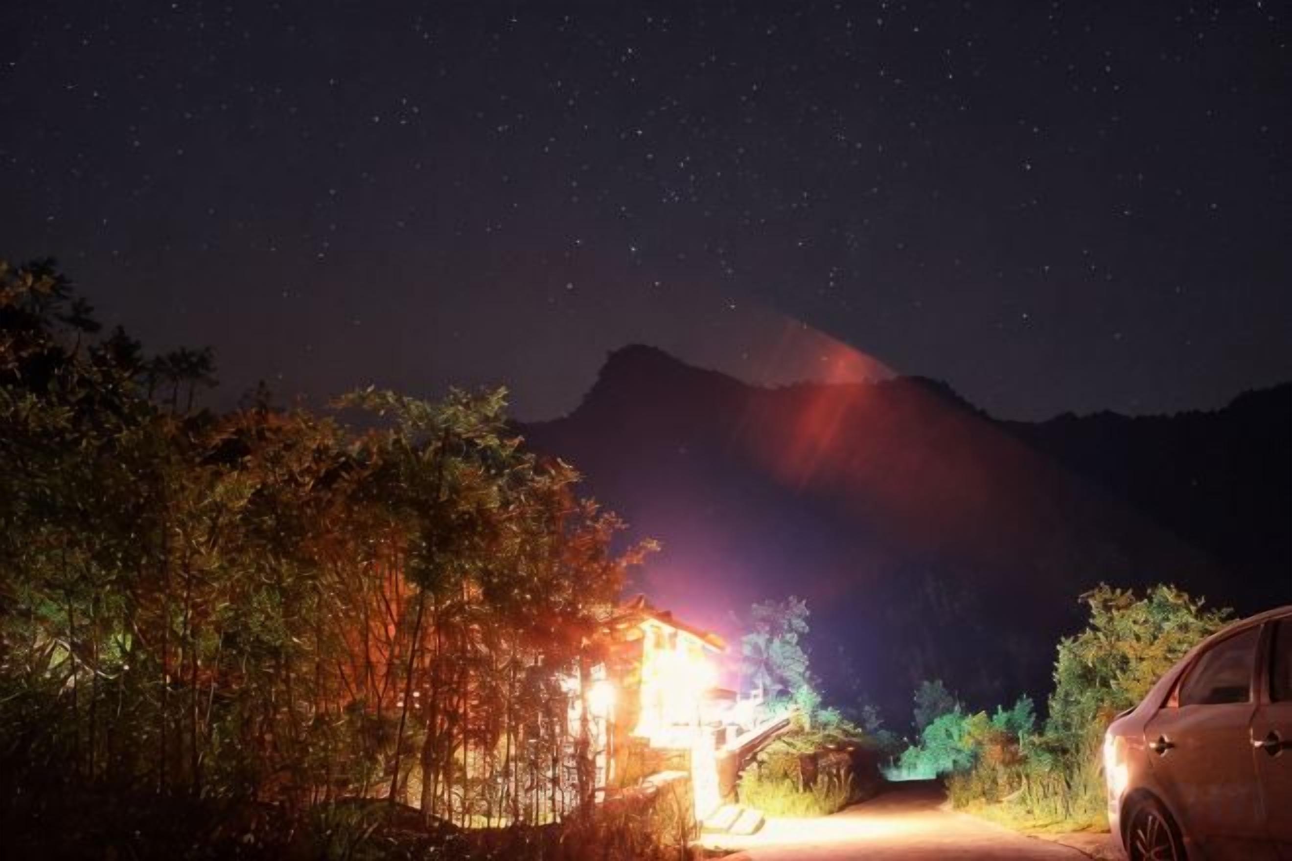 Escena nocturna con una montaña al fondo, un edificio iluminado y un coche aparcado.