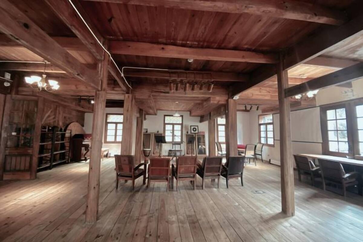 Une pièce spacieuse dotée de planchers et de poutres en bois, comprenant un espace salon central avec des chaises et des tables, ainsi que de grandes fenêtres laissant entrer la lumière naturelle.