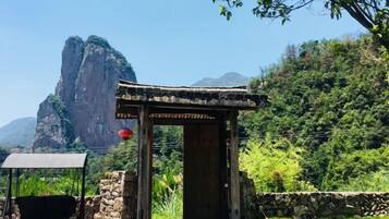 A traditional wooden structure with a stone base, a swing, and a red lantern, set against a backdrop of lush greenery and a prominent rock formation.