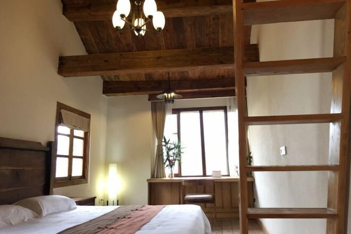 Quarto com cabeceira de madeira, cama com roupa de cama branca, escrivaninha com cadeira e uma janela grande com cortinas.