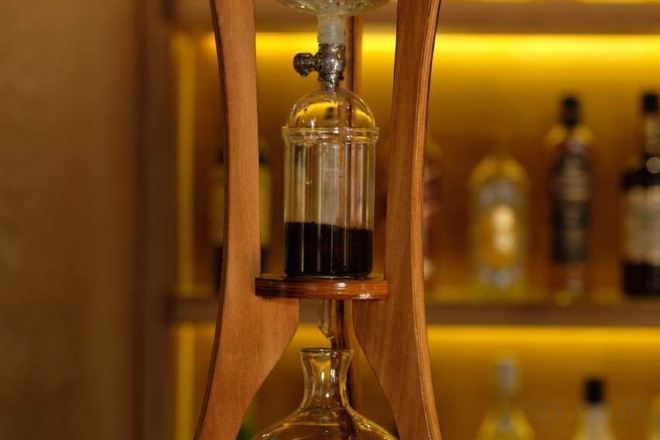 Una estructura de madera con un recipiente de vidrio, posiblemente un aparato de destilación, y un estante con botellas al fondo.