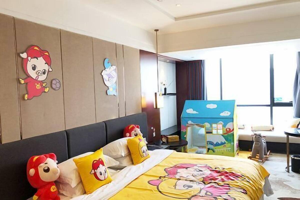 Una habitación de hotel con cama, almohadas, una mesita de noche, cómoda y una ventana, además de una casita de juguete.