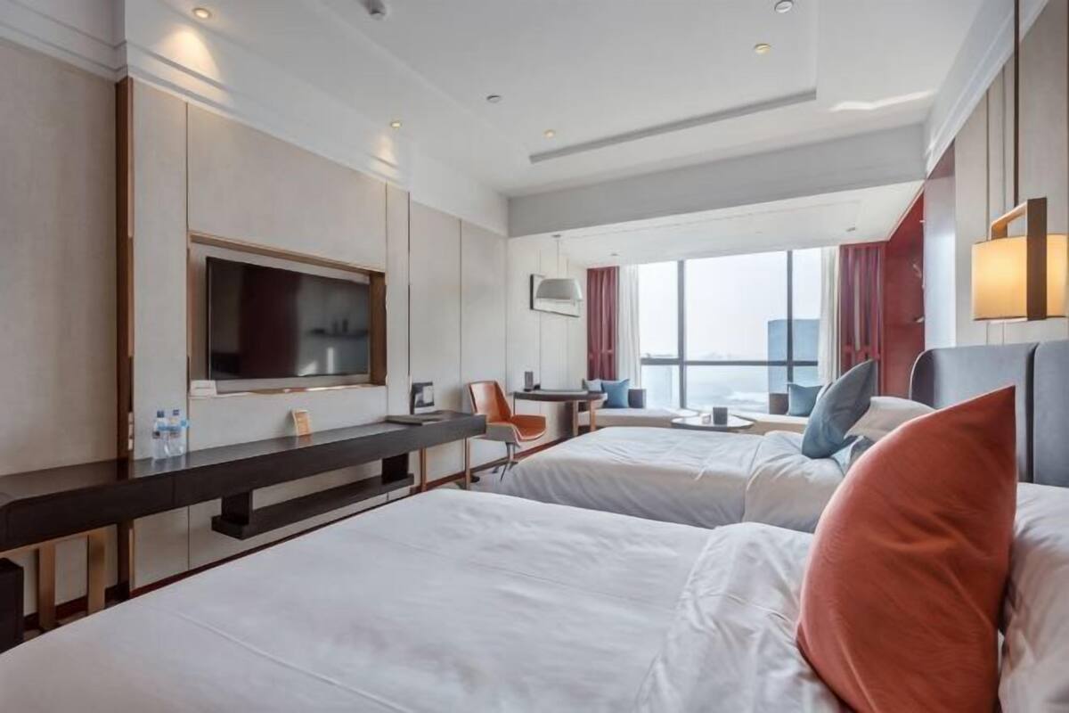 Habitación de hotel con dos camas, un escritorio, un televisor y un ventanal grande.