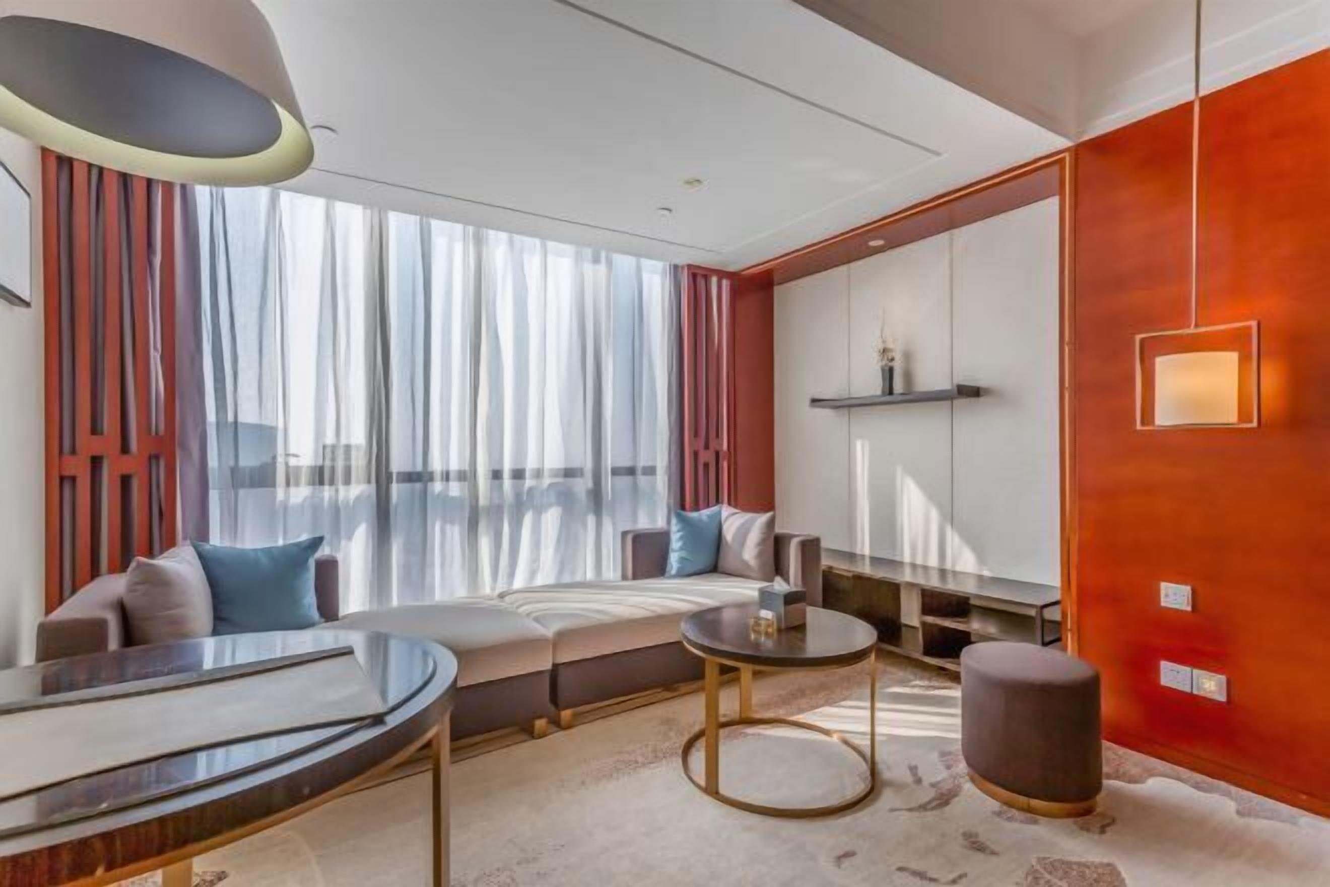 Une chambre d’hôtel moderne, équipée d’un canapé, d’une table ronde, d’un petit tabouret et d’une grande fenêtre avec des rideaux.