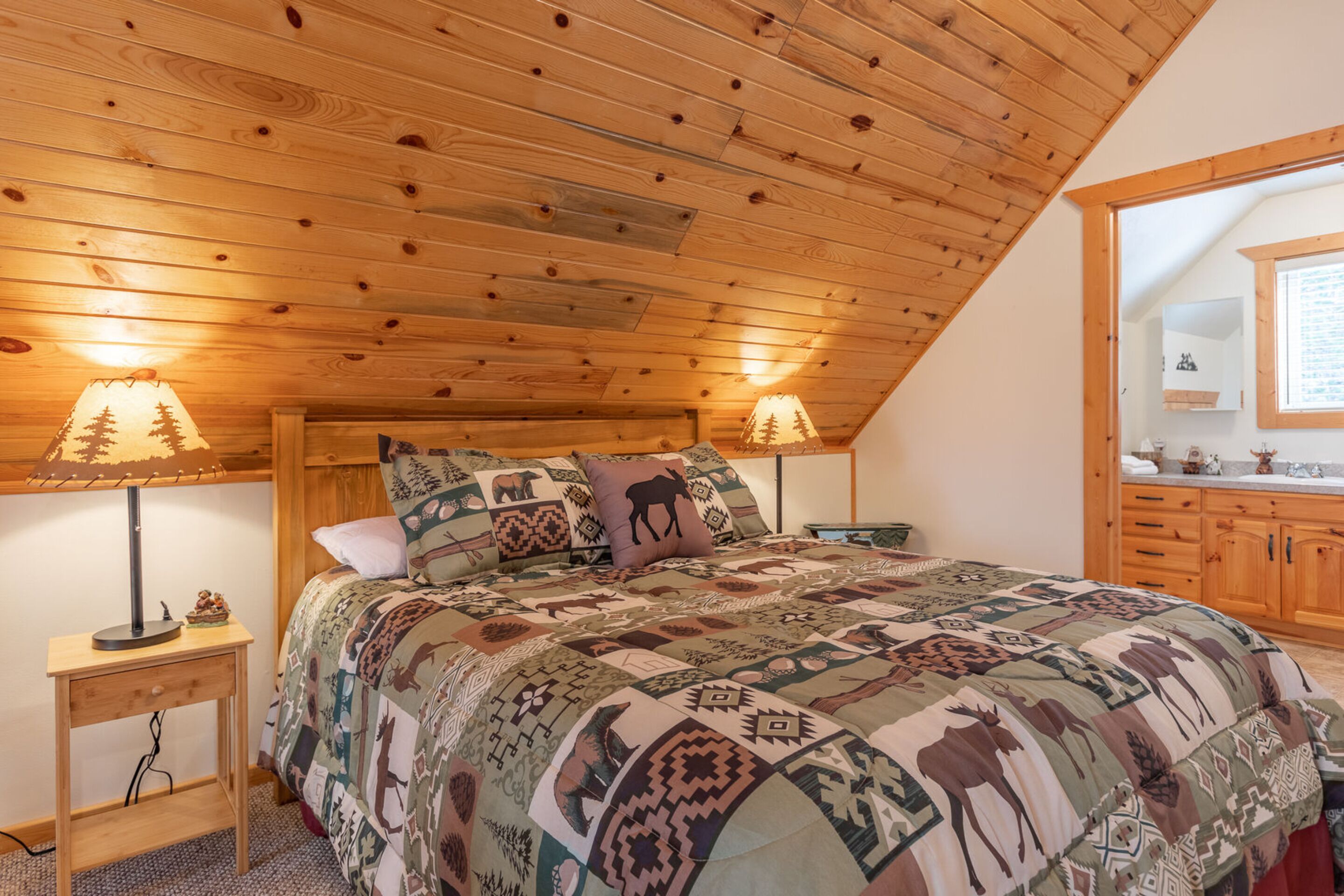 Un dormitorio con techo de madera, una cama con colcha estampada, dos lámparas de noche y una mesita de noche de madera.