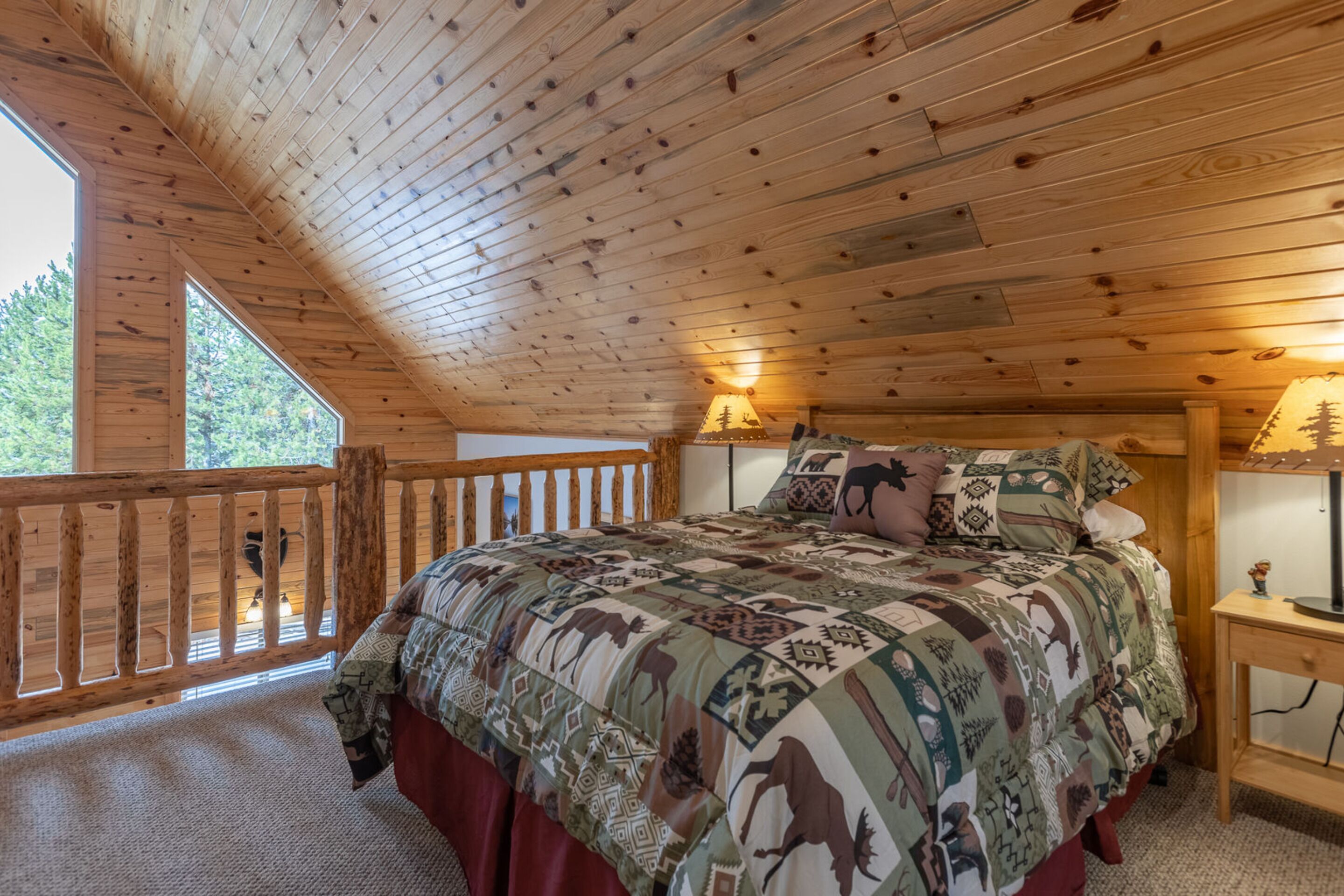 A wooden cabin bedroom with a large bed, a nightstand, and a window overlooking greenery.