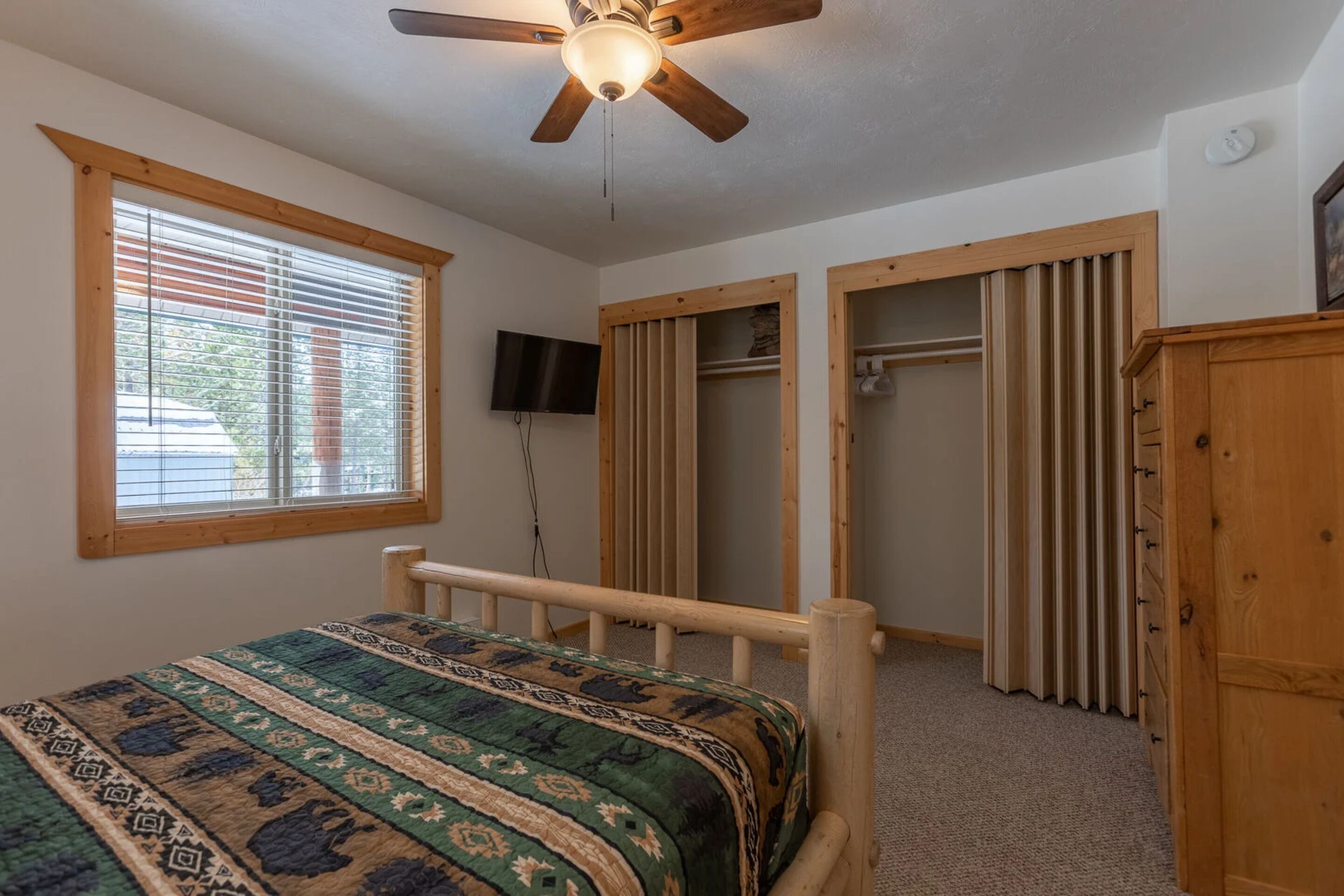 A bedroom with a bed, a ceiling fan, a window with blinds, a TV, and a wooden wardrobe.