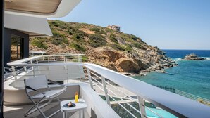 A balcony with white chairs and a table, overlooking a rocky coastline with clear blue waters and a building on a hill. - Made By The Sea Villas 12 Bdrm (Malevizi)