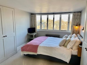 A bedroom with a bed, a television on a small table, a window with curtains, and a radiator.