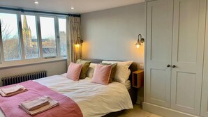 A bedroom with a bed, bedside lamps, a radiator, a wardrobe, and a window with a view of a building.