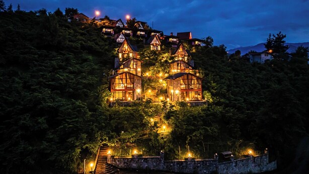 A large, illuminated castle-like structure surrounded by dense forest, with a stone staircase leading up to it. - GAFULLUK TATİL KÖYÜ (Arakli)