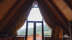 An attic room with a stone wall, a wooden sloped ceiling, and a large window with curtains. - GAFULLUK TATİL KÖYÜ (Arakli)