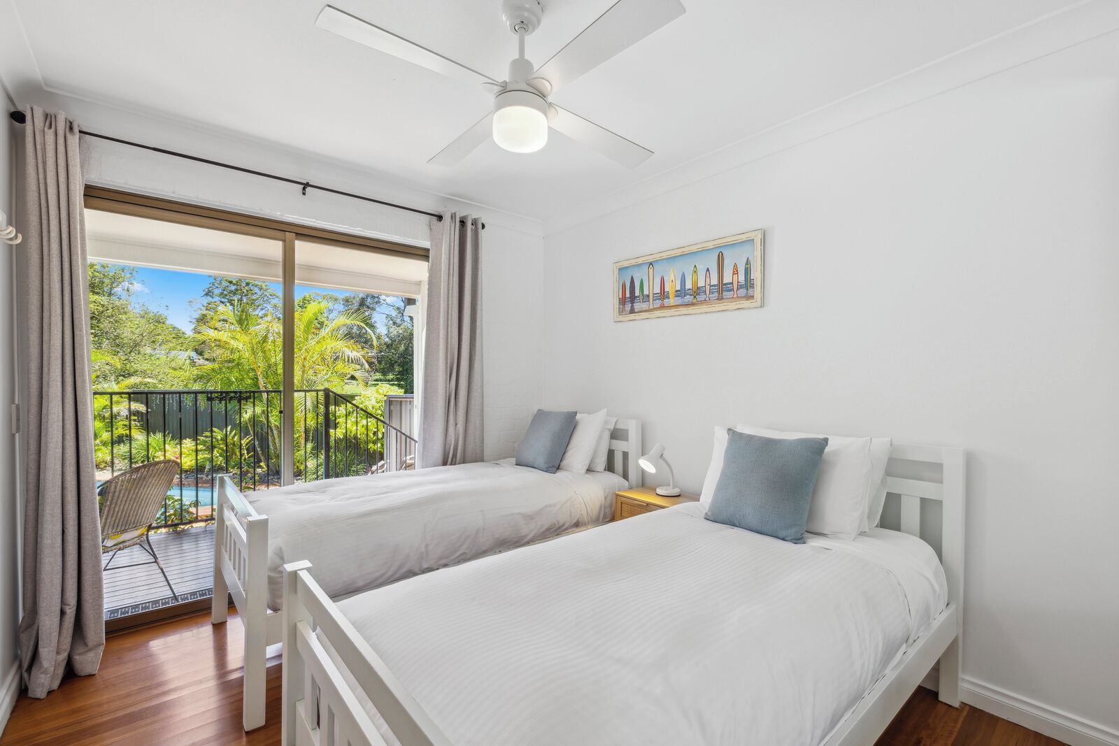 A bedroom with two beds, a ceiling fan, a painting, and a balcony with a chair and a pool.