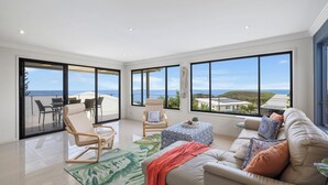 Ein geräumiges Wohnzimmer mit einem großen Fenster, das Blick auf das Meer und die Hügel bietet. Es gibt ein Sofa, einen Sessel und einen kleinen Tisch mit einem Teppich.