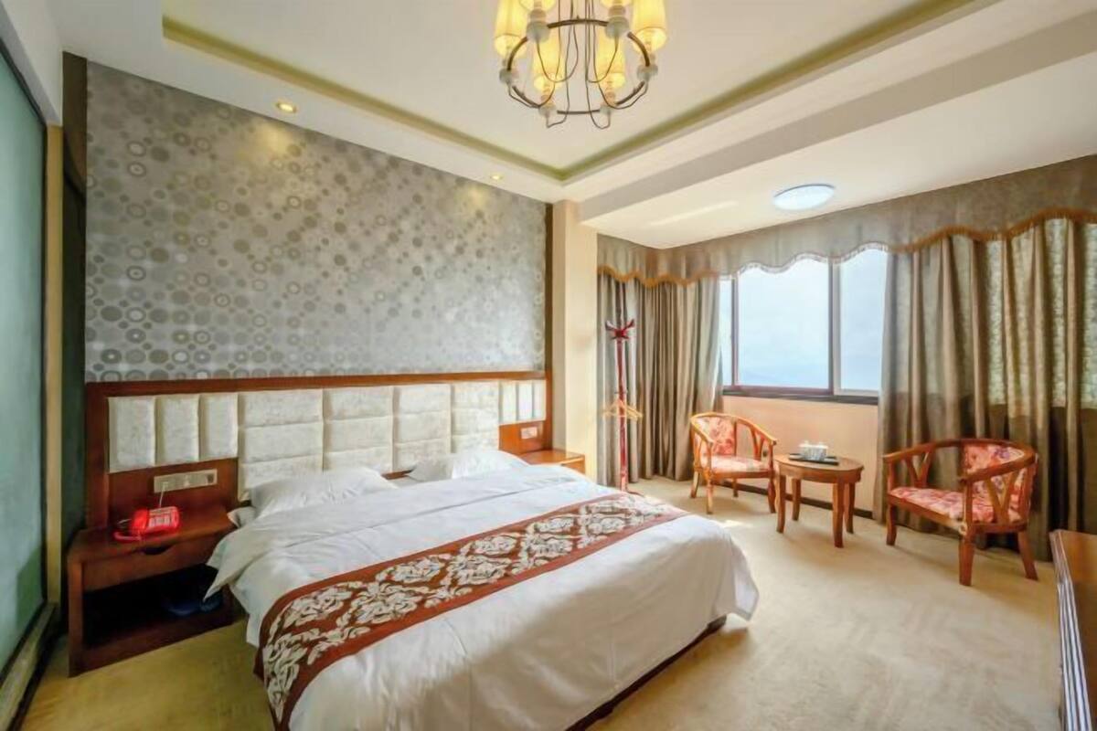 Quarto de hotel com uma cama grande, uma mesa de cabeceira com luminária, uma cadeira, uma mesinha e uma janela com cortinas.