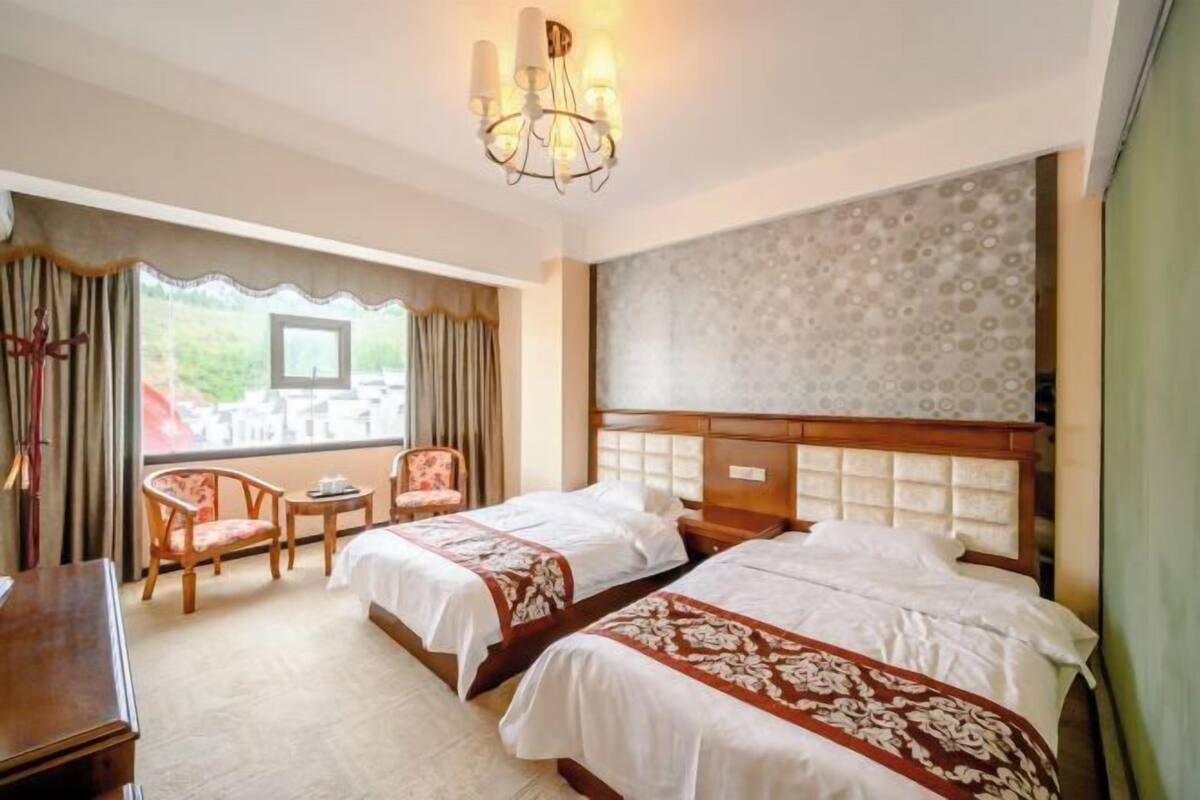 Quarto de hotel com duas camas, uma escrivaninha, uma cadeira, um candelabro e vista para o verde lá fora.