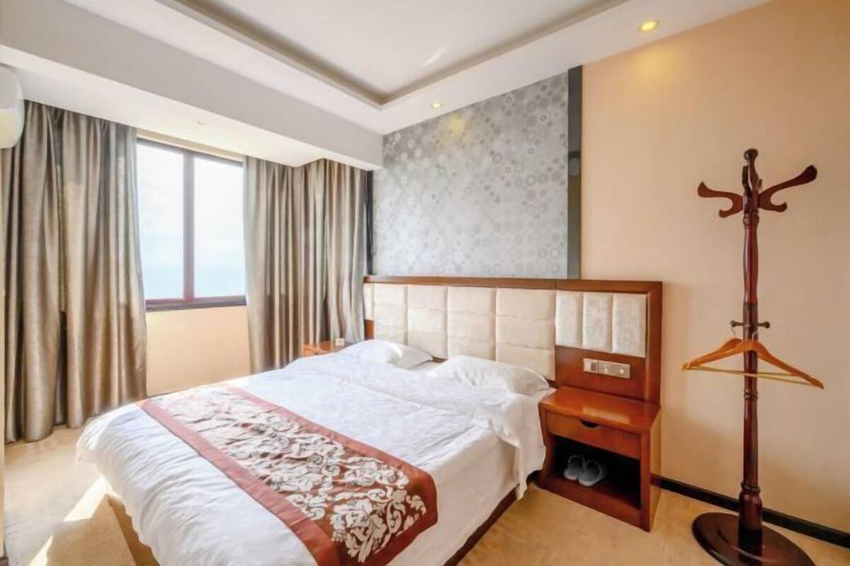 Quarto de hotel com uma cama grande, uma mesa de cabeceira de madeira, um porta-roupas e uma janela com cortinas.