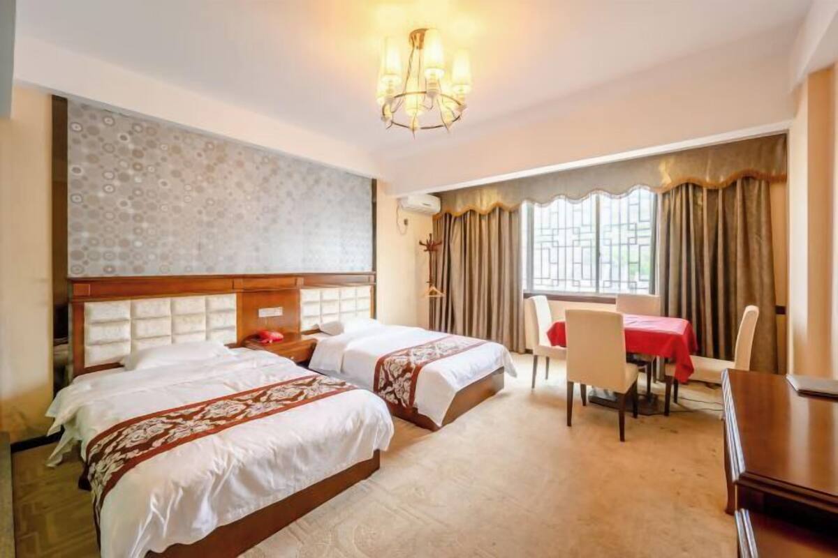 Quarto de hotel com duas camas, uma mesa de jantar, cadeiras, um candelabro e uma janela com cortinas.