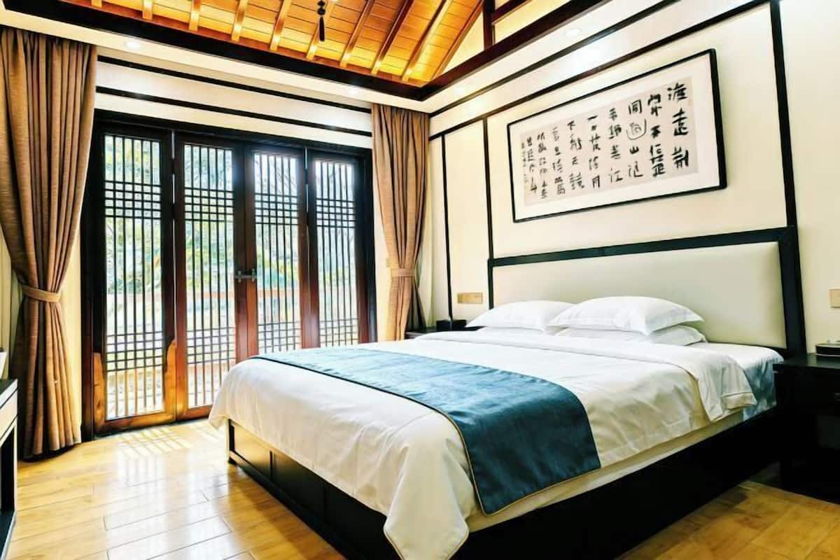 Une chambre d’hôtel avec un lit, des rideaux, une grande fenêtre donnant sur un beau paysage et un mur orné de calligraphies.