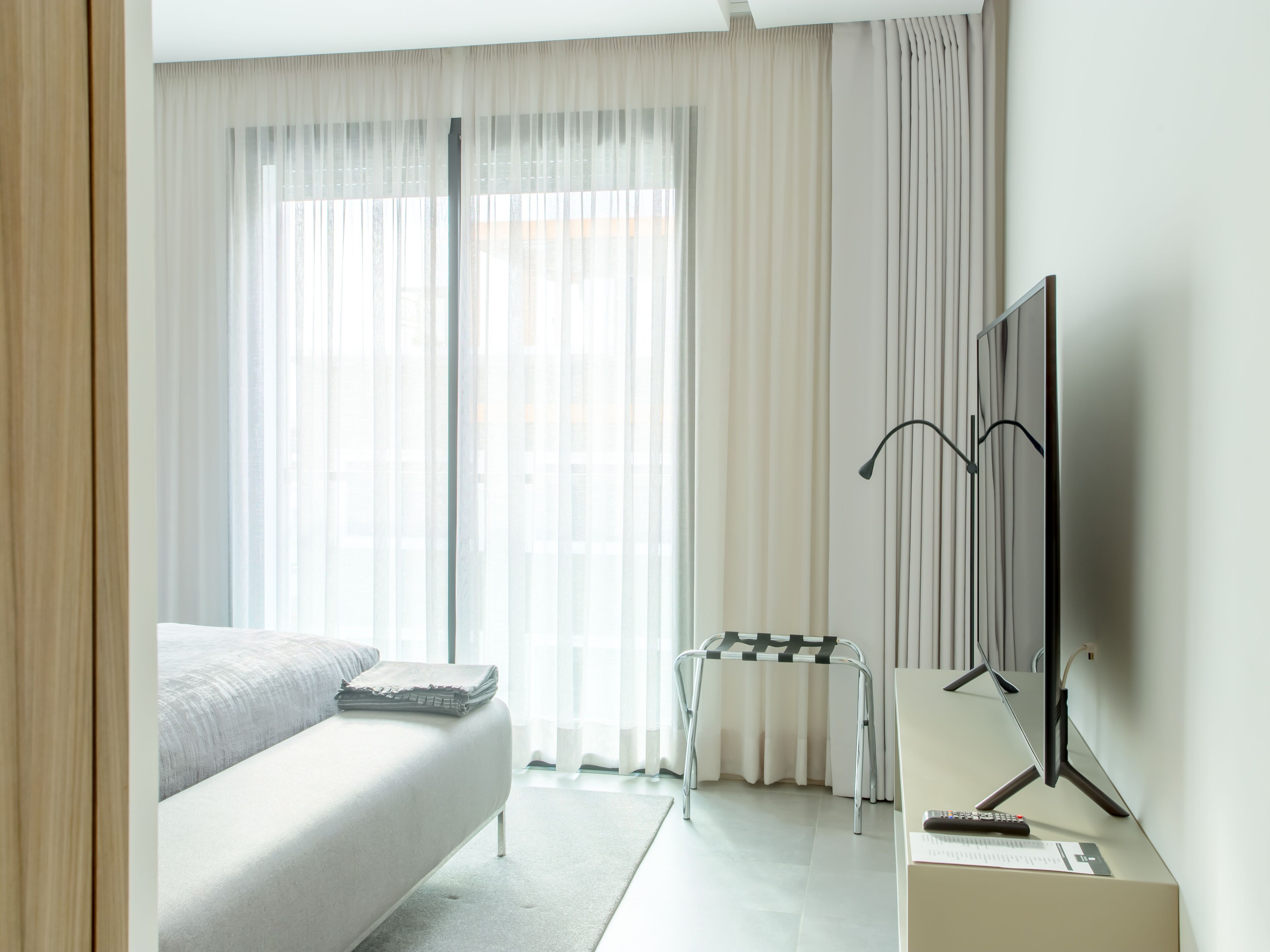 A modern hotel room with a bed, a desk with a computer, and a large window with sheer curtains.