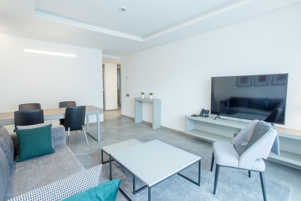 A modern living room with a sofa, two armchairs, a coffee table, a TV, and a desk with a phone.