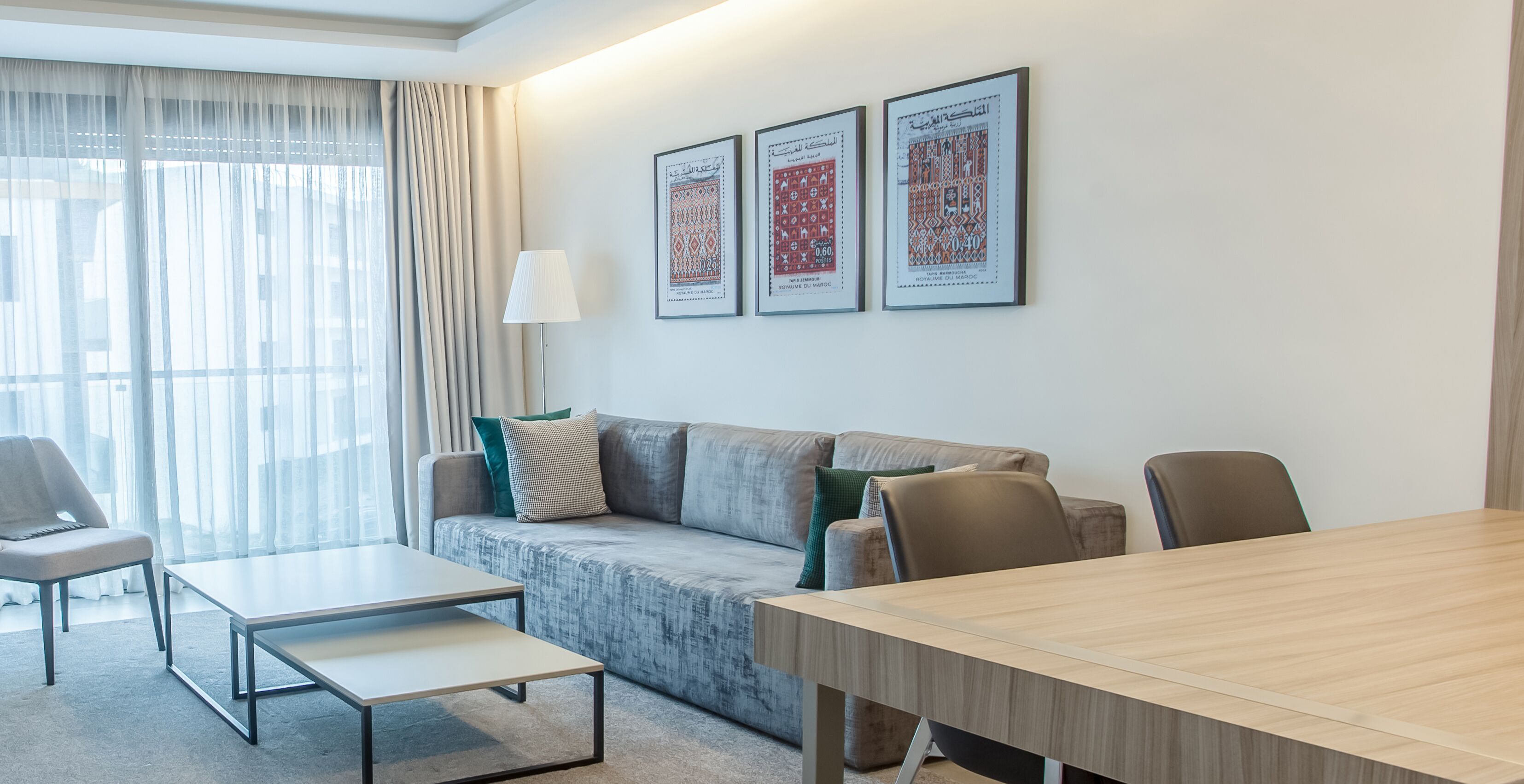 A modern hotel room with a grey sofa, a small table, a wooden dining table, and a lamp. There are three framed pictures on the wall.