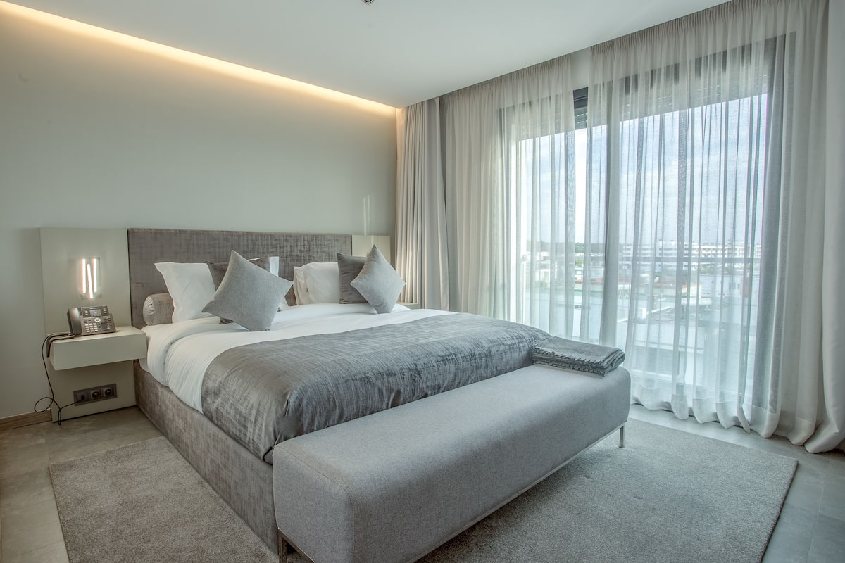 A modern hotel room with a large bed, a bedside table with a lamp, and a view of the city through sheer curtains.